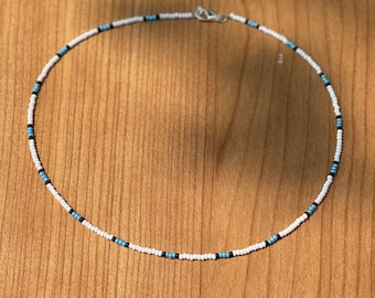 May include: A beaded necklace with a white base, accented with small turquoise and black beads. The necklace is circular and has a silver clasp. The beads are evenly spaced, creating a simple yet stylish accessory.