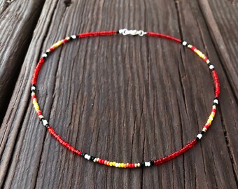 May include: A beaded necklace with a silver clasp. The necklace features small, colorful beads in red, black, white, and yellow. The necklace is displayed on a rustic, wooden surface.