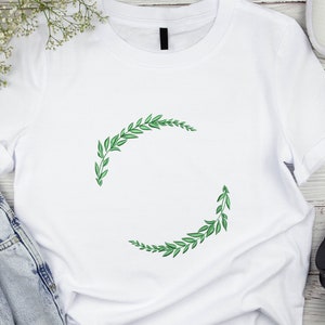 May include: White t-shirt featuring a green leafy wreath design. The wreath is made of two half circles of leaves.