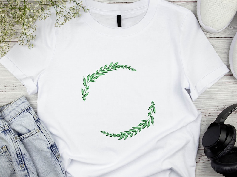 May include: White t-shirt featuring a green leafy wreath design. The wreath is made of two half circles of leaves.