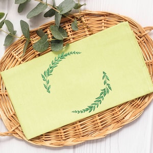 May include: A light green tea towel with a green embroidered wreath design. The towel is folded and placed in a wicker basket.