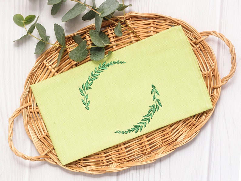 May include: A light green tea towel with a green embroidered wreath design. The towel is folded and placed in a wicker basket.