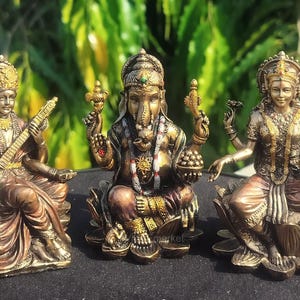 Puede incluir: Tres estatuas de color bronce de deidades hindúes. Las estatuas representan a Saraswati tocando un instrumento de cuerda, a Ganesha con cabeza de elefante y a Lakshmi sosteniendo una olla. Cada figura está adornada con joyas detalladas y sentada sobre una base de flor de loto.