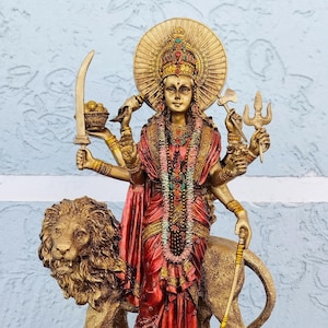 May include: A detailed golden statue of a deity with multiple arms, wearing a red and gold dress, and a halo. The deity stands on a lion and holds various objects, including a sword and a trident. The statue has intricate details and is set against a light blue background.
