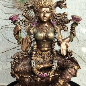 May include: A bronze-colored statue of a seated deity, adorned with intricate details. The figure holds lotus flowers and wears a crown and garland. A peacock feather is visible in the background. The statue is a representation of a goddess.