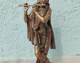 Puede incluir: Estatua de bronce de una figura tocando una flauta. La figura lleva una corona, joyas y ropa drapeada en tonos rojos y dorados. La estatua se alza sobre una base redonda adornada con flores moradas.