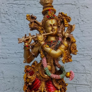 Op de afbeelding: Een gouden beeld van een godheid die fluit speelt. Het beeld heeft ingewikkelde details, waaronder een sierlijke hoofdtooi, sieraden en een rood en goud kledingstuk. Een bloemenslinger hangt om de nek. De achtergrond is een lichtgrijze bakstenen muur.