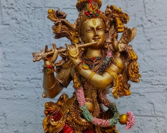 Puede incluir: Estatua dorada de una deidad tocando la flauta. La estatua presenta detalles intrincados, incluyendo un tocado ornamentado, joyas y una prenda roja y dorada. Una guirnalda de flores cuelga alrededor del cuello. El fondo es una pared de ladrillos gris claro.