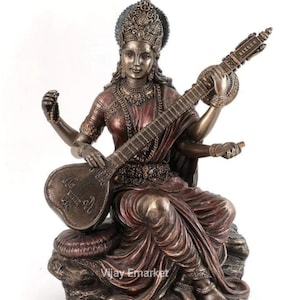 Puede incluir: Una estatua de bronce de Saraswati, la diosa hindú del conocimiento, la música y el arte. Se la representa tocando un veena, un instrumento de cuerda, y está adornada con intrincadas joyas y una corona. La estatua está colocada sobre una base de roca y tiene un acabado rico y multicolor.