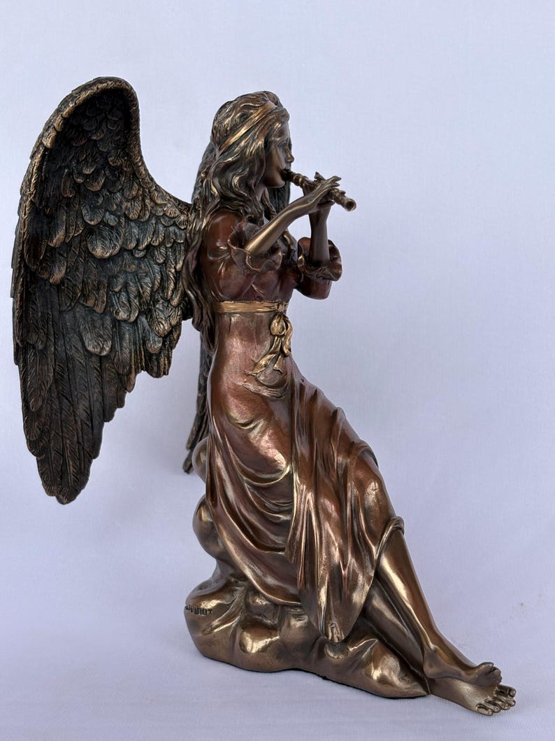 May include: A bronze-toned sculpture of a seated angel with large, detailed wings, wearing a flowing dress with a tied waist. The figure is depicted playing a small flute, with long hair and bare feet, resting upon a cloud-like base structure.