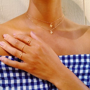 Puede incluir: Una persona lleva un collar de oro con dos colgantes de iniciales, una 'M' y una 'R'. La persona también lleva tres anillos de oro en su dedo.
