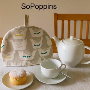 May include: A white tea set with a teapot, teacup, saucer, and a cake on a plate. A patterned tea cosy with bird designs covers the teapot. The text "SoPoppins" is at the top.