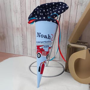 Könnte beinhalten: Ein blauer und weißer, kegelförmiger Schultüte mit einem roten Band. Die Tüte ist mit Sternen, einem Feuerwehrauto und dem Namen "Noah" mit dem Text "Kindergartenkind 2024" verziert.