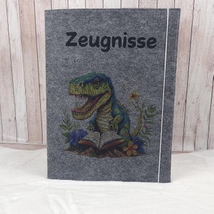 Könnte beinhalten: Grauer Filzordner mit einem weißen Reißverschluss und dem Wort "Zeugnisse" auf der Vorderseite. Auf der Vorderseite ist eine bunte Illustration eines Dinosauriers zu sehen, der ein Buch liest.