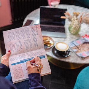 Op de afbeelding: Een vrouw zit aan een tafel met een laptop, een kop koffie en een tijdschrift. Ze schrijft in een planner met de tekst "Be Prepared" op de kaft. De planner heeft een roze en blauw ontwerp.
