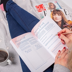 Op de afbeelding: Een roze en wit dagboek met de woorden "Know your worth" en "Comparing Yourself" op de omslag. Het dagboek ligt open op een pagina met prompts voor zelfreflectie en het stellen van doelen. Het dagboek ligt op een wit, pluche deken met de benen van een persoon in blauwe jeans zichtbaar op de achtergrond.