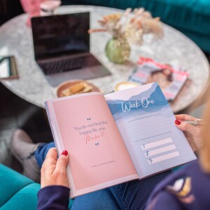 Op de afbeelding: Een open dagboek met een roze omslag en blauwe pagina's. Het dagboek heeft een citaat op de omslag dat luidt: "Je vindt het gelukkige leven niet, je cre&euml;ert het." Het dagboek is open op een pagina met de titel "Week &eacute;&eacute;n" en bevat een lijst met prompts voor de week.