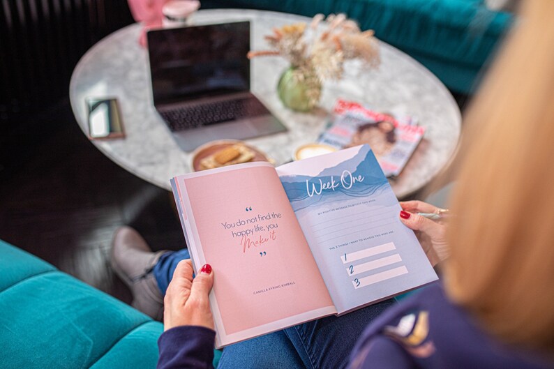 Op de afbeelding: Een open dagboek met een roze omslag en blauwe pagina's. Het dagboek heeft een citaat op de omslag dat luidt: "Je vindt het gelukkige leven niet, je cre&euml;ert het." Het dagboek is open op een pagina met de titel "Week &eacute;&eacute;n" en bevat een lijst met prompts voor de week.