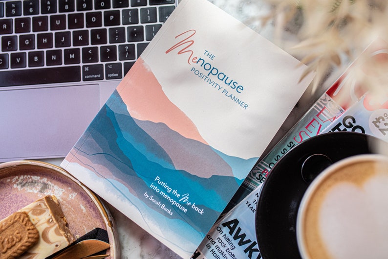Op de afbeelding: Een wit boek met een blauw en roze aquarelontwerp op de omslag. Het boek is getiteld "The Menopause Positivity Planner: Putting the "Me" Back into Menopause" door Sarah Banks.