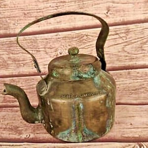 Puede incluir: Una tetera de latón vintage con un acabado desgastado y un asa desgastada. La tetera tiene un cuerpo redondeado y un pico en el lateral.