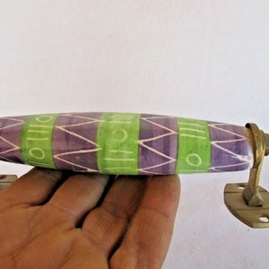 May include: A decorative cabinet pull with a ceramic handle featuring a purple and green geometric design. The handle is mounted on brass hardware.