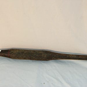 May include: A weathered, wooden rolling pin with a cylindrical center and tapered ends. The wood has a dark, aged appearance with visible grain and a reddish-brown band near the center. A small white label is attached.