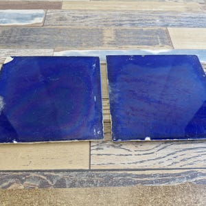 May include: Two square, cobalt blue tiles with glossy surfaces. The tiles have slight imperfections and are set against a wooden plank background. The tiles are likely for home decor or construction.