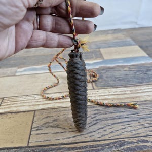 May include: A dark brown, cone-shaped pendant with a textured surface, hanging from a multicoloured twisted cord. The pendant is set against a wooden plank background.