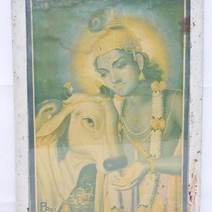 May include: A framed vintage print of a Hindu deity, Krishna, with a white cow. Krishna is depicted with a blue body, a yellow crown, and a garland of flowers. The print is set against a green and yellow background.