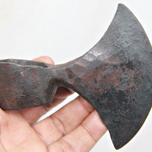 Op de afbeelding: Een roestige, handgesmede bijl met een gebogen blad en een afgeronde steel. De bijl is gemaakt van metaal en heeft een donkerbruine patina.
