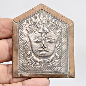Può includere: Una placca in argento e rame con un'immagine in rilievo di una divinità con una corona e un baffo. La placca è rettangolare con una parte superiore a punta e un bordo decorativo.