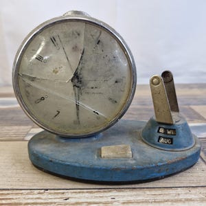 May include: An antique blue desk clock with a broken glass face and a calendar feature. The clock has a silver-colored bezel and a round face with Roman numerals. The calendar displays the day and month.