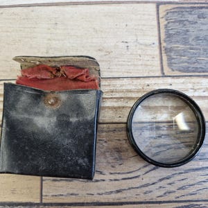 Puede incluir: Un estuche de cuero negro con interior rojo y una lente redonda y transparente con borde negro. El estuche está abierto, revelando el forro rojo. La lente está junto al estuche sobre una superficie de madera.