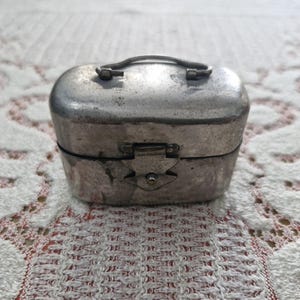May include: A small, oval-shaped silver-toned metal container with a smooth, rounded lid and a central handle. The box features a front-facing latch mechanism for closure, with a weathered, metallic finish showing signs of age and use.