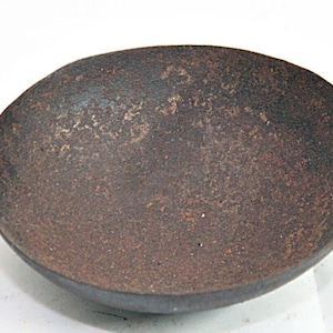 Op de afbeelding: Een kleine, roestige, ijzeren pan met twee handvatten. De pan is rond en ondiep, met een licht gebogen bodem. De handvatten zijn van ijzer en zijn bevestigd aan de zijkanten van de pan.