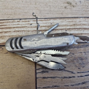 Puede incluir: Una multiherramienta plateada vintage con varios implementos extendidos, incluyendo un sacacorchos, un cuchillo y unas tijeras. La herramienta tiene un mango texturizado con tres hendiduras curvas. El metal muestra signos de edad y uso.