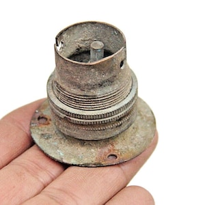 Puede incluir: Un casquillo de bombilla vintage de latón con una base roscada y un punto de contacto central. El casquillo está desgastado y tiene una pátina.