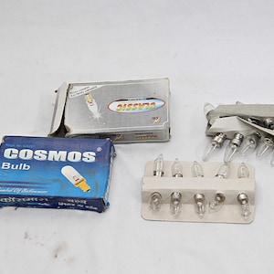 Könnte beinhalten: Eine Auswahl kleiner Glühbirnen und ihre Verpackungen. Zwei Kartons sind sichtbar, einer blau mit der Aufschrift "COSMOS Bulb" und ein silberner Karton mit der Aufschrift "CLASSIC". Mehrere Glühbirnen sind in weißen Haltern angeordnet.