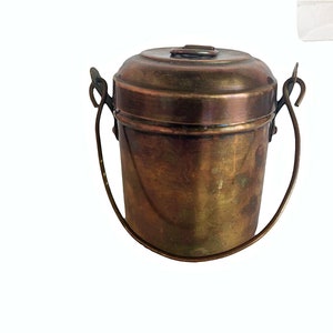 May include: A small, round, copper container with a lid and a handle. The container has a worn, aged look.