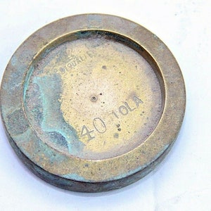 Op de afbeelding: Een rond messinggewicht met een verweerde, groene patina. Het gewicht is gemarkeerd met "40 TOLA" en "QUALITEIT".