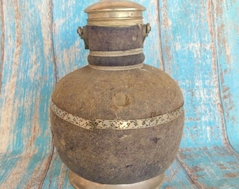 Könnte beinhalten: Eine braune Lederwasserflasche mit einem silbernen Deckel und einem dekorativen Band. Die Flasche hat ein rustikales, vintage Aussehen.