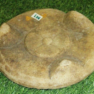 May include: A round, light brown stone with a raised, four-leaf clover design in the center. The stone has four raised points on the underside, suggesting it was used as a base or stand.