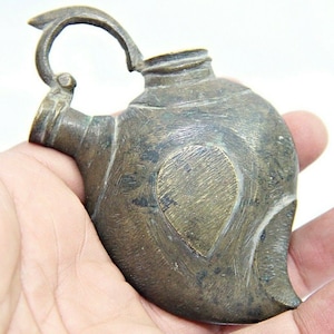 Può includere: Una piccola lampada ad olio antica in ottone con un corpo a forma di cuore e un manico. La lampada ha un beccuccio su un lato e una piccola apertura rotonda sulla parte superiore.