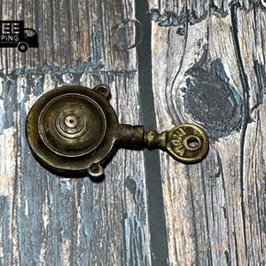 Op de afbeelding: Een messing sleutel met een ronde, decoratieve kop. De sleutel heeft een cirkelvormig ontwerp met concentrische cirkels en een klein gaatje in het midden. De sleutel ligt op een houten oppervlak.