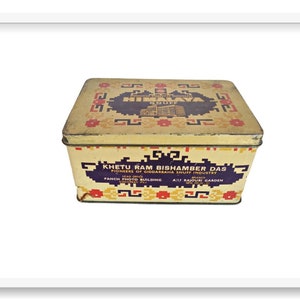 Puede incluir: Una caja de hojalata vintage con un diseño amarillo, rojo y azul. La caja está etiquetada con "KHETU RAM BISHAMBER DAS" y "PIONEERS OF GIDDARBAHA SNUFF INDUSTRY".