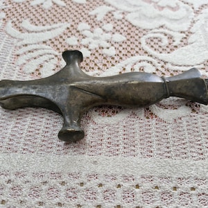Op de afbeelding: Een dof messing metalen voorwerp in de vorm van een kruis, met een langwerpig centraal lichaam, een uitlopend uiteinde en een kleine uitstekende vierkante punt. Het oppervlak vertoont tekenen van ouderdom, oxidatie en een matte afwerking.