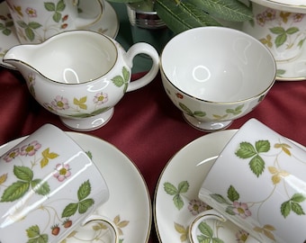 Puede incluir: Un juego de seis tazas de té y platillos blancos con un patrón floral de fresas y hojas. Las tazas y los platillos tienen un borde dorado. El juego está dispuesto sobre una superficie roja con una rama de bayas rojas.