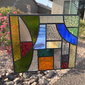 May include: A rectangular stained glass panel featuring a geometric arrangement of multicolored glass pieces. The design includes various textures and hues, such as deep green, vibrant orange, cobalt blue, red, and clear textured glass, all held together by dark metal lead lines.