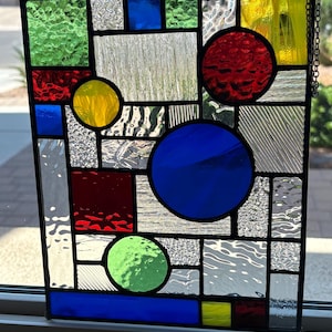 May include: A stained glass panel with a geometric design. The panel features various shapes in red, yellow, green, and blue, with clear and textured glass sections. A black metal frame and chain complete the piece.