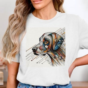 May include: White t-shirt featuring a colorful, geometric illustration of a dog's head in profile. The artwork uses lines and shapes in shades of blue, orange, yellow, and brown. The dog's eye is visible, and the design has a modern, artistic feel.